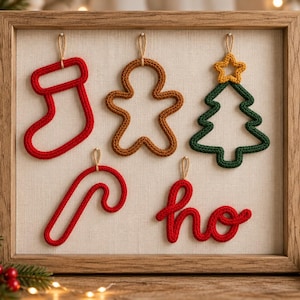 May include: A wooden frame containing five hanging knitted ornaments: a red stocking, a brown gingerbread person, a green Christmas tree with a yellow star, a red candy cane, and the red cursive text ho, all suspended by small twine loops.