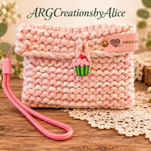 May include: A light pink and white hand-knitted pouch with a wrist strap, featuring a cupcake-shaped button closure and a pink leather tag embossed with a heart and the text ARGCreationsbyAlice. The pouch has a textured knit pattern.