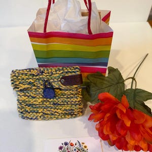 Puede incluir: Una pequeña bolsa tejida a mano con hilo jaspeado en amarillo, azul y verde, con un cierre de botón azul oscuro y una etiqueta de cuero marrón. Se acompaña de una bolsa de regalo a rayas de colores, una flor artificial naranja y una tarjeta blanca con el texto ARG Creations By Alice.