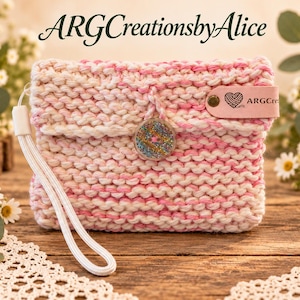 Könnte beinhalten: Eine handgestrickte Tasche aus weißem und rosafarbenem Garn mit einer weißen Handschlaufe. Die Tasche verfügt über einen runden, glitzernden Knopfverschluss und ein hellrosa Lederetikett mit Herz- und Garnsymbol sowie dem Text ARGCreationsbyAlice.