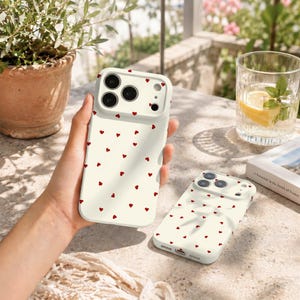 May include: Two off-white smartphone cases featuring a repeating pattern of small red hearts. The cases have a raised camera cutout area with circular openings for lenses and a flash, designed to fit a modern smartphone with a triple camera system.