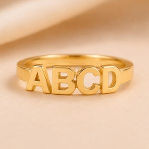 Pode incluir: Um anel de tom dourado com as letras maiúsculas A, B, C e D dispostas em linha ao longo do topo da banda. As letras estão integradas na estrutura do anel, criando uma superfície metálica contínua e polida com um acabamento liso.