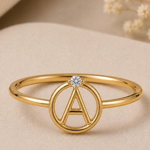 May include: A gold ring featuring a circular frame with the letter A centered inside. A small, clear, round-cut gemstone is set at the top of the circular frame where it meets the thin, polished gold band, creating a delicate and refined appearance.