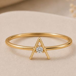 以下が含まれることがあります： 中央に三角形のアクセントがある細いゴールドリング。三角形は2本の角度のついた金属バーが先端で合わさる形で構成されており、その中央には小さく透明なラウンドカットの宝石がしっかりとセットされています。
