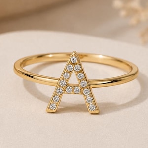 May include: A gold-toned ring featuring a capital letter A as the centrepiece. The letter A is embellished with small, clear, round-cut stones set along its surface. The band of the ring is thin, smooth, and circular, maintaining a consistent gold-coloured finish throughout the entire piece.