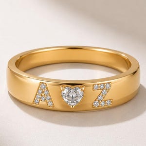Puede incluir: Un anillo de banda dorada con la letra A, una piedra en forma de corazón y la letra Z. Las letras A y Z están incrustadas con pequeñas piedras claras, y la piedra en forma de corazón está situada en el centro entre ambas letras.