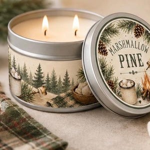 May include: A round silver metal candle tin with two lit wicks. The label features a forest scene with pine trees, a campfire, and mugs of marshmallows. The lid displays the text MARSHMALLOW PINE surrounded by pine branches, pinecones, and a campfire illustration.