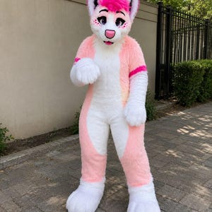 May include: A full-body fursuit costume featuring a white and light pink colour scheme with a plush texture. The head piece has large pointed ears with bright pink tips, a pink nose, and purple eyes. The suit includes large white paws.