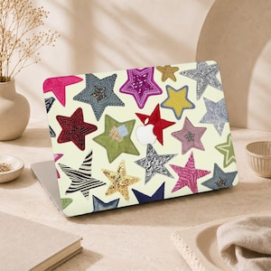 May include: A laptop skin featuring a cream-coloured background covered in a variety of star-shaped patches. The stars display diverse textures and patterns, including glitter, zebra stripes, denim, metallic silver, gold, and embroidered fabric designs.