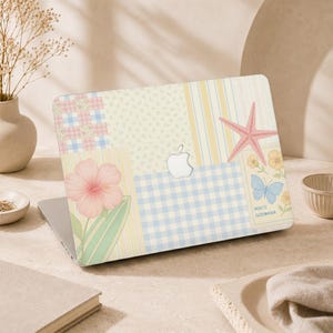 Op de afbeelding: Een laptopsticker met een patchworkontwerp in pastelgeel, blauw en roze, inclusief ruitjes, strepen en bloemenprints. Het ontwerp bevat een roze zeester, een roze hibiscusbloem, een blauwe vlinder en de tekst POSTE GODWANA.