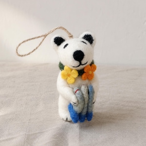 Könnte beinhalten: Ein weißes Filz-Bärenornament mit schwarzen Augen und Nase, das ein grünes Halsband mit gelben und orangefarbenen Blumen trägt. Der Bär hält drei kleine graue Fische mit blauen Schwänzen und hat eine dünne braune Schnurschlaufe am Kopf.