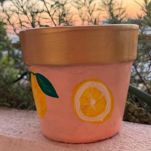 Op de afbeelding: Een kleine terracotta plantenpot geschilderd in een zachte perzikkleur met een metallic gouden rand. De buitenkant is voorzien van handgeschilderde gele citroenschijfjes en een groen bladontwerp. De pot heeft een taps toelopende vorm.