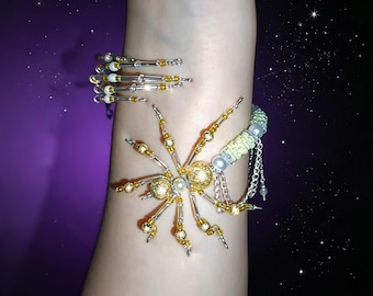 May include: A beaded bracelet featuring a central spider motif crafted from gold and silver-toned beads, accented with small white pearls. The band consists of a coiled wire design with alternating gold and silver beads, finished with a woven section.