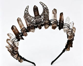 May include: A headband featuring a series of raw, dark-toned quartz crystal points secured with black wire. A silver-toned metal crescent moon charm with intricate filigree cutouts is positioned at the center, layered over the central crystal point.