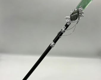 May include: A hair stick featuring a long, smooth black shaft adorned with a silver-toned metal wire wrap. The wire is shaped into delicate leaf patterns and secures a translucent green crystal point at the top, alongside a small dark stone.