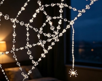 May include: A decorative spider web made of clear, faceted crystal-like beads strung together to form a geometric web pattern. A single beaded spider hangs from the bottom of the web, and the entire structure is illuminated by small white lights.