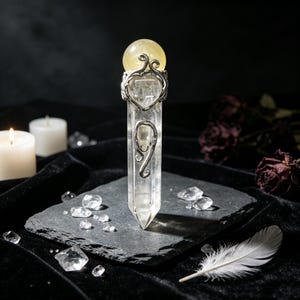 May include: A clear, hexagonal quartz crystal wand topped with a yellow spherical stone. The crystal is encased in a silver-toned metal filigree featuring a heart-shaped design and swirling patterns, standing vertically on a dark, textured stone base.
