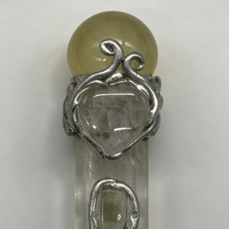 May include: A vertical wand-like object featuring a clear, cylindrical crystal base topped with a smooth, pale yellow spherical stone. A silver-toned metal wire wrap forms a heart shape around a clear crystal and decorative swirls on the shaft.