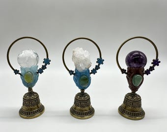 May include: Three decorative stands featuring a brass-toned bell base, a central holder with a green stone, and a circular wire frame adorned with small blue or purple flowers. The stands hold a clear sphere, a white crystal cluster, and a purple sphere.