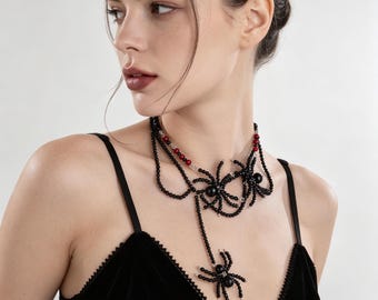 May include: A multi-strand beaded necklace featuring black and red spherical beads, accented with two black spider-shaped charms. The necklace includes a long, dangling strand ending in a third spider charm, all crafted with small black beads.