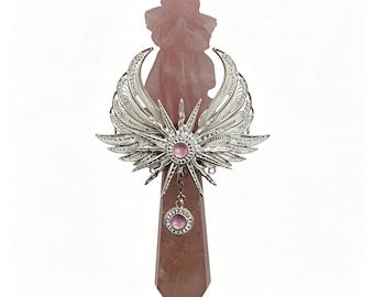 Puede incluir: Una figura de ángel tallada en cuarzo rosa montada sobre una punta de cristal de cuarzo rosa. La pieza está adornada con un adorno de metal plateado con alas, un diseño de rayos de sol y dos pequeños cabujones rosas circulares.