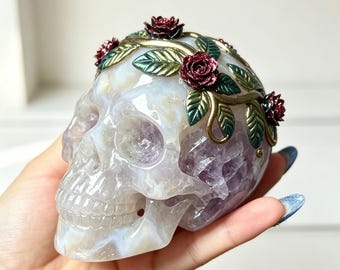 May include: A carved amethyst skull sculpture featuring a decorative metal vine crown. The crown is adorned with metallic green leaves and small, deep red glittery roses that wrap around the top of the translucent purple and white stone skull.