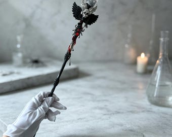 May include: A black decorative wand featuring a clear crystal sphere at the top, accented by a silver skull and black wings. The handle is adorned with a black spider, red beads, and intricate dark detailing, creating a gothic-inspired aesthetic.