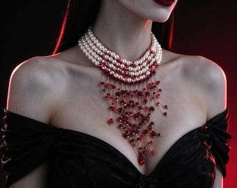 May include: A multi-strand choker necklace featuring white pearls accented with red splatters. A cascading fringe of translucent red beads and teardrop-shaped charms hangs from the center, creating a layered, dangling effect against the neck.