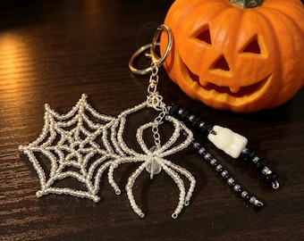 May include: A silver-toned metal keychain featuring a beaded spider web and a beaded spider made of clear, translucent beads. Attached are two dangling strands of black beads and a single white tooth-shaped charm on a silver split ring.