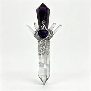 Puede incluir: Un objeto vertical en forma de varita con una base larga de cristal de cuarzo transparente rematada con una punta de amatista de color p&uacute;rpura oscuro. Un alambre de metal plateado envuelve el centro, sujetando una peque&ntilde;a piedra p&uacute;rpura en forma de coraz&oacute;n.