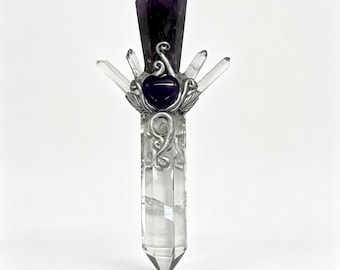 May include: A vertical wand-like object featuring a long clear quartz crystal base topped with a dark purple amethyst point. Silver-toned metal wire wraps around the center, holding a small purple heart-shaped stone and four smaller clear crystals.