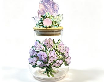 May include: A clear glass jar featuring a decorative lid topped with a large, translucent purple crystal and smaller clear crystals set in green leaves. The jar body is adorned with a sculpted green vine design holding clusters of light purple stones.