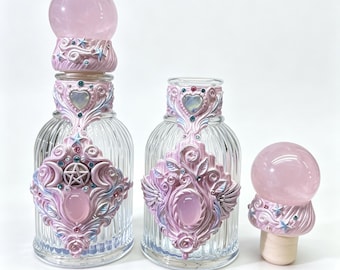 May include: Two clear glass bottles with vertical ribbing, each featuring ornate pink and blue sculpted clay embellishments, heart-shaped accents, and a pentacle symbol. One bottle is topped with a spherical pink crystal stopper.
