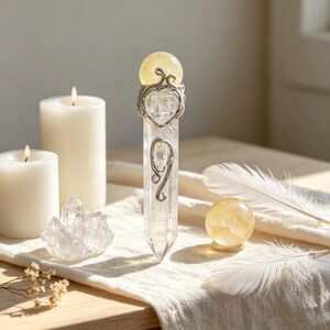 May include: A clear quartz crystal wand topped with a yellow citrine sphere, featuring silver-toned metal filigree accents. Beside it are two white pillar candles, a small clear quartz cluster, a yellow citrine sphere, and two white feathers.