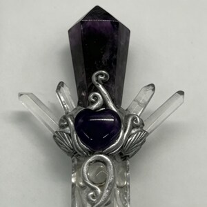Puede incluir: Una varita decorativa con una punta de amatista de color morado oscuro en la parte superior, una piedra central morada en forma de coraz&oacute;n y cuatro puntas de cuarzo transparente hacia afuera. Las piedras est&aacute;n en un marco de metal plateado.