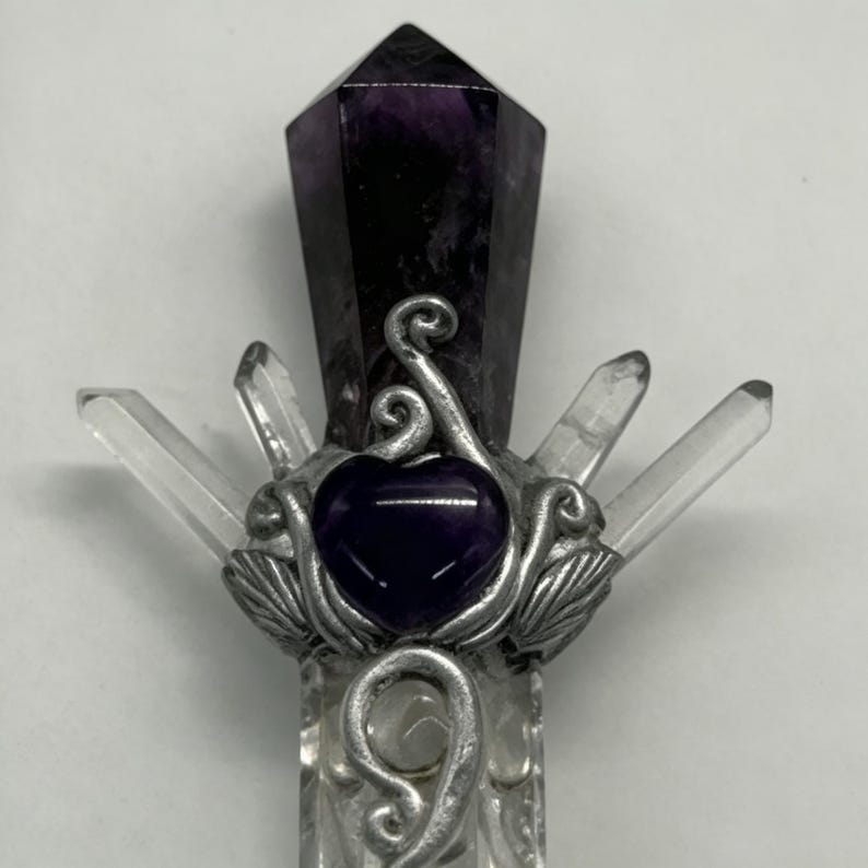 Puede incluir: Una varita decorativa con una punta de amatista de color morado oscuro en la parte superior, una piedra central morada en forma de coraz&oacute;n y cuatro puntas de cuarzo transparente hacia afuera. Las piedras est&aacute;n en un marco de metal plateado.