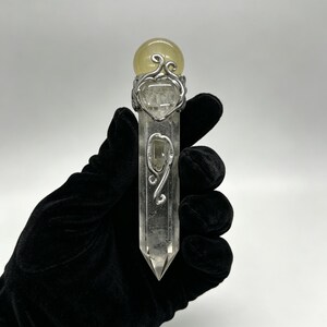 May include: A clear, hexagonal quartz crystal wand topped with a smooth, pale yellow spherical stone. The crystal is wrapped in decorative silver-toned wire filigree that forms intricate, swirling patterns along the upper and middle sections.