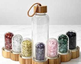 May include: A clear glass water bottle with a bamboo lid and a brown cord loop, surrounded by six smaller glass capsules with bamboo bases. Each capsule contains different colored tumbled stones: red, clear, yellow, purple, pink, green, and black.