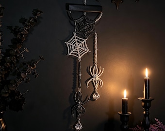 May include: A hanging wall decoration featuring two crocheted spider webs in black and silver, two beaded spider figures, and two clear faceted crystal pendants suspended from a horizontal black wooden bar by silver chains.