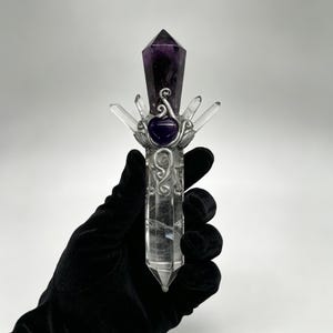 Puede incluir: Una varita de cristal de cuarzo transparente con una punta de amatista de color p&uacute;rpura oscuro en la parte superior. La varita est&aacute; adornada con alambre de metal plateado, una peque&ntilde;a piedra de amatista en forma de coraz&oacute;n y cuatro puntas.