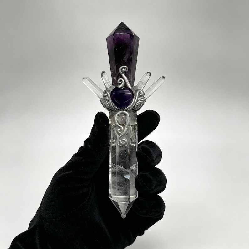 Puede incluir: Una varita de cristal de cuarzo transparente con una punta de amatista de color p&uacute;rpura oscuro en la parte superior. La varita est&aacute; adornada con alambre de metal plateado, una peque&ntilde;a piedra de amatista en forma de coraz&oacute;n y cuatro puntas.