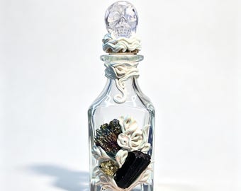 May include: A clear glass bottle featuring a transparent skull-shaped stopper, adorned with white sculpted clay swirls, a dark iridescent mineral cluster, a black tourmaline crystal, and a small piece of gold-colored pyrite at the base.