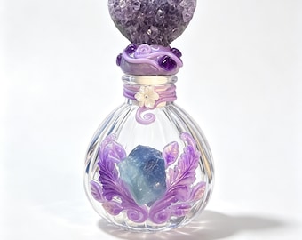 May include: A clear glass bottle with vertical ribbing, featuring a heart-shaped amethyst crystal stopper. The neck is wrapped in purple clay with a white flower accent. The front displays a blue crystal surrounded by purple clay leaves.
