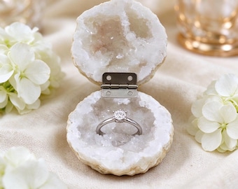 May include: A silver ring with a single round-cut clear stone sits inside an open, hollowed-out white geode ring box. The geode features a rough, crystalline interior and is connected by a small, rectangular silver-colored metal hinge.