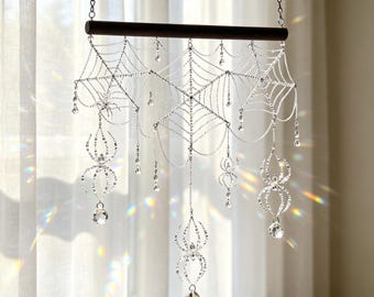 May include: A hanging wall decoration featuring a dark horizontal wooden rod with silver chains forming a spider web pattern. Several dangling silver spider charms and clear faceted crystal beads hang from the web, catching light to create prisms.