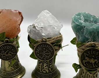 May include: Three decorative stands featuring raw crystals in peach, clear, and green. Each stand is wrapped in twine and green leaves, with a bronze-toned metal charm attached. The charms are embossed with the zodiac signs Virgo, Sagittarius, and Aries.