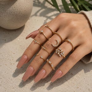 May include: A set of gold-toned rings worn on fingers, featuring various designs including a crescent moon, a leaf-shaped cluster with clear stones, small faux pearls, and thin bands with embedded crystals. The rings are arranged across multiple fingers.