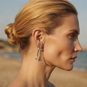 May include: A silver ear cuff featuring a smooth, continuous metal band that loops around the outer ear cartilage. The design consists of two parallel, curved lines that extend downward, creating a sleek, polished, and minimalist metallic accessory.