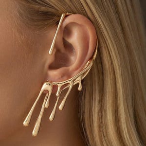 May include: A gold-toned ear cuff featuring a fluid, melted design that extends along the outer edge of the ear. The piece consists of a smooth, metallic structure with multiple elongated, drip-like segments hanging downward from the earlobe.