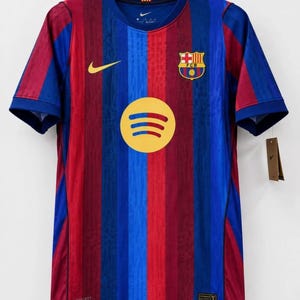 May include: A short-sleeved football shirt featuring vertical blue and dark red stripes with a subtle tonal pattern. The shirt displays a yellow Spotify logo in the centre, a yellow Nike swoosh on the right chest, and the FC Barcelona crest on the left.